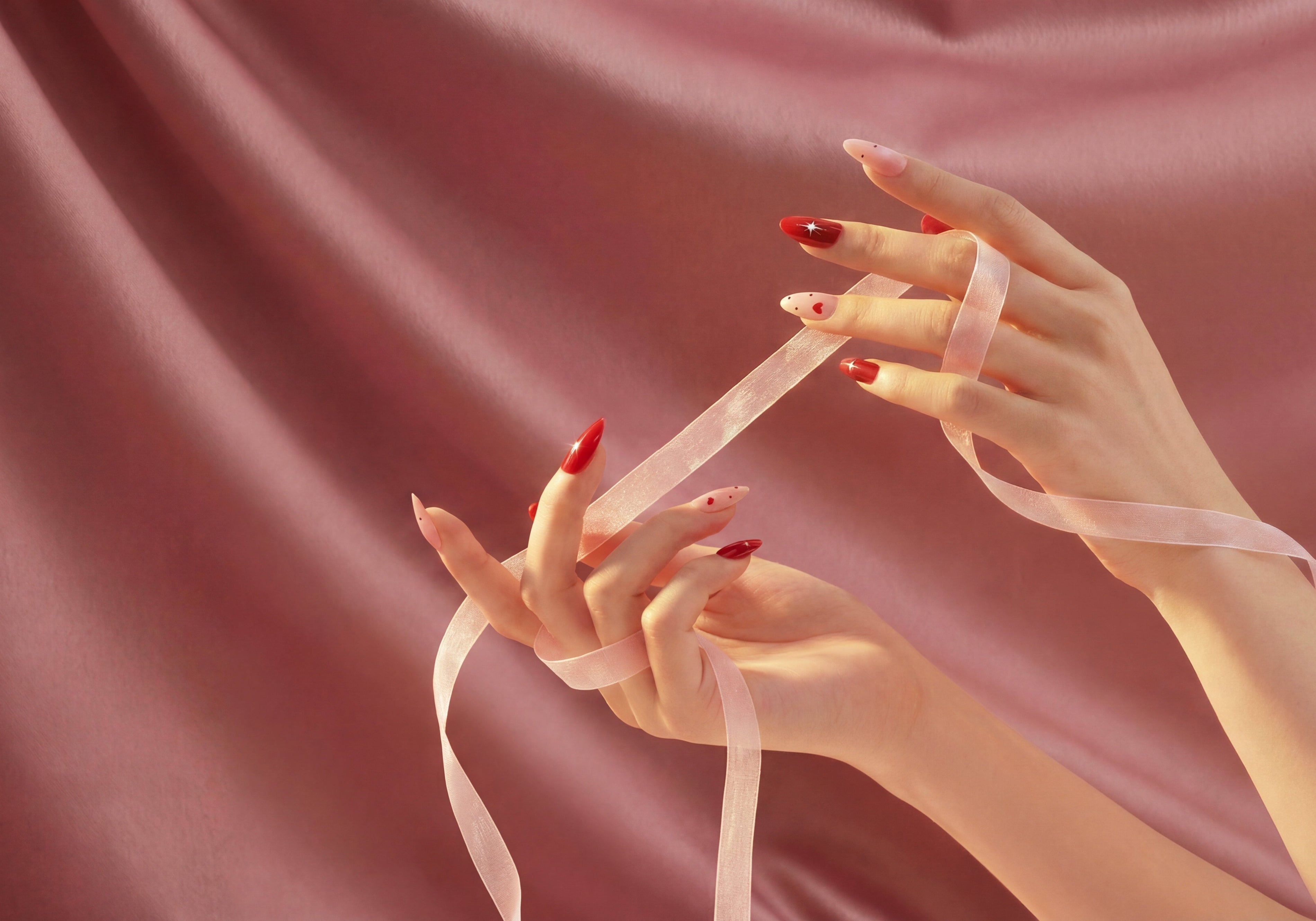 Hand holding pink ribbon with red and nude almond-shaped press-on nails, pink satin background
