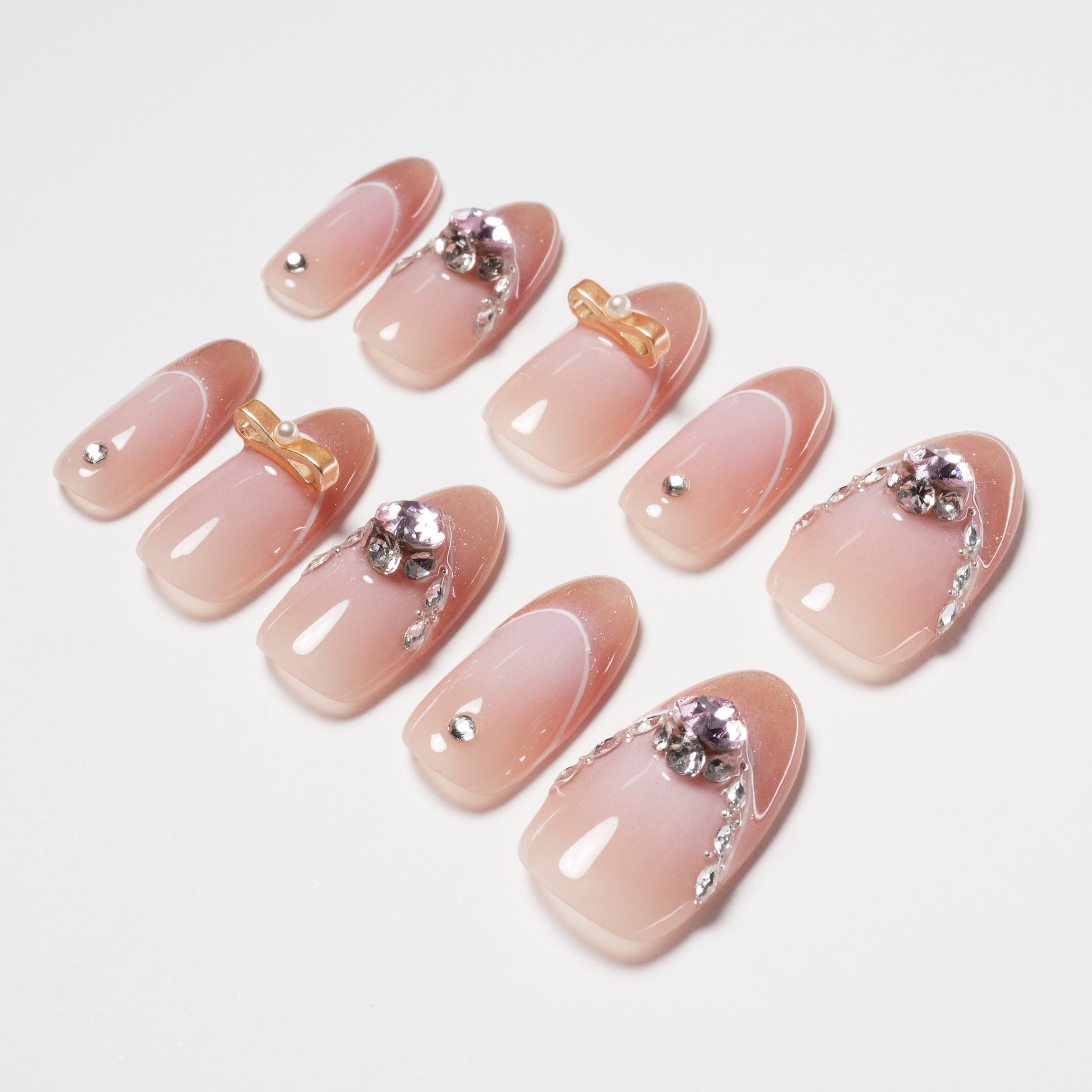 Handmade pink press-on nails with rhinestones, gold bows, and pearl accents, arranged on white background.