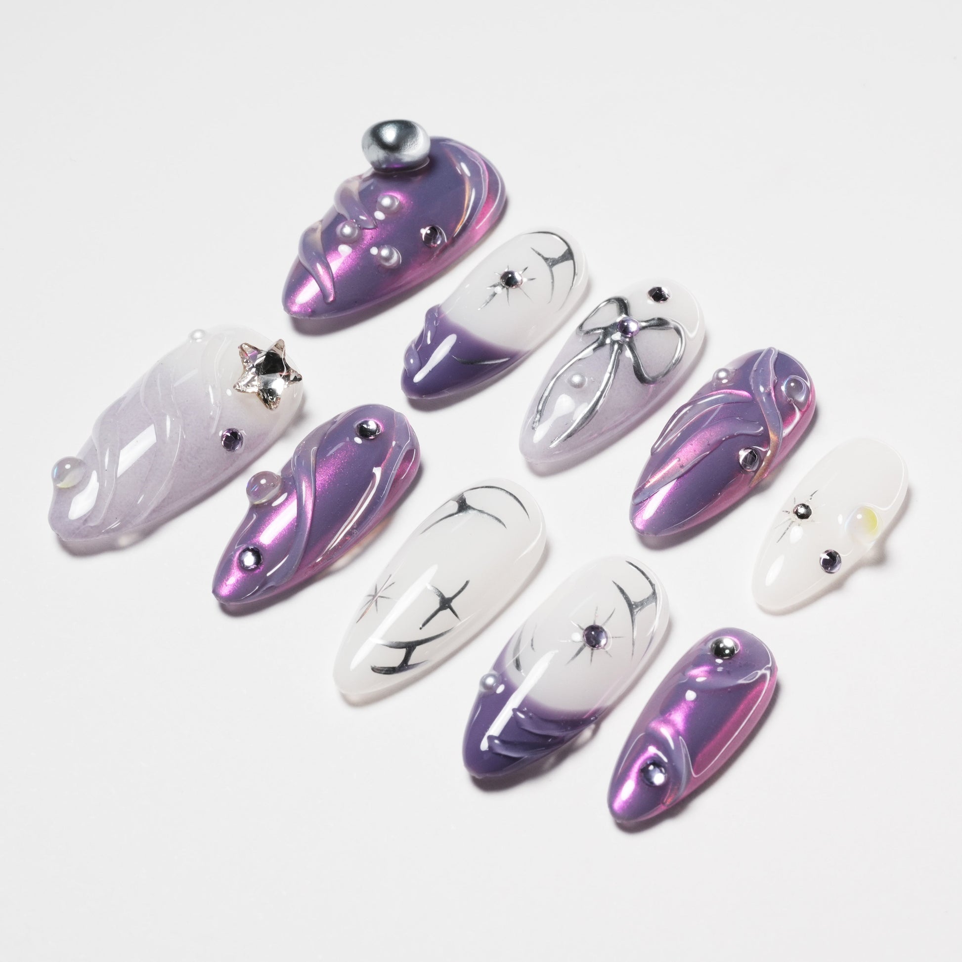 Purple and white press-on nails decorated with silver gems, pearl accents, and black abstract designs