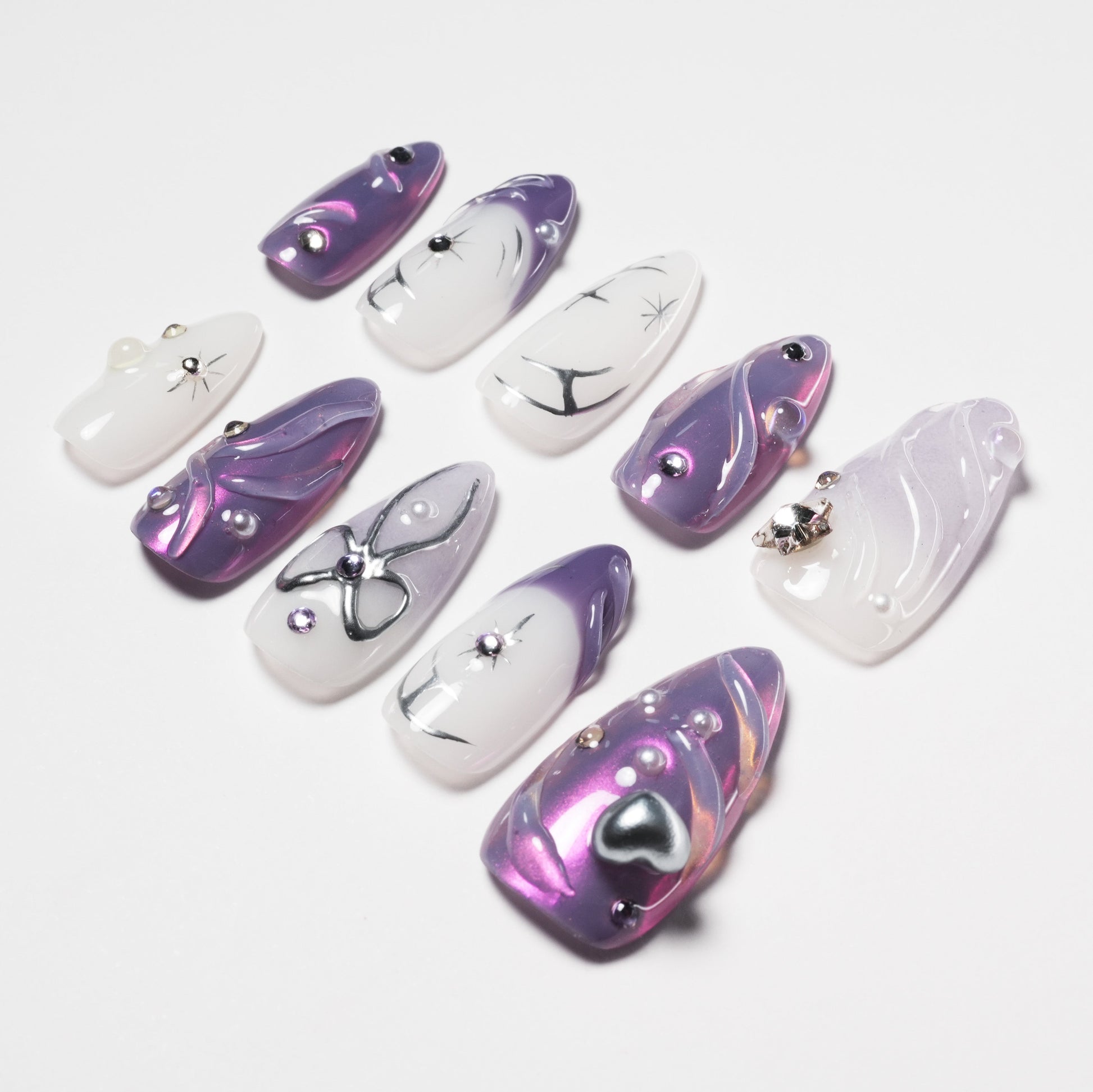 Set of purple and white artificial nails with metallic and pearl embellishments on white background