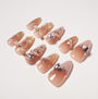 Set of glossy nude gradient press-on nails with gold tips, bows, and pink heart rhinestone accents