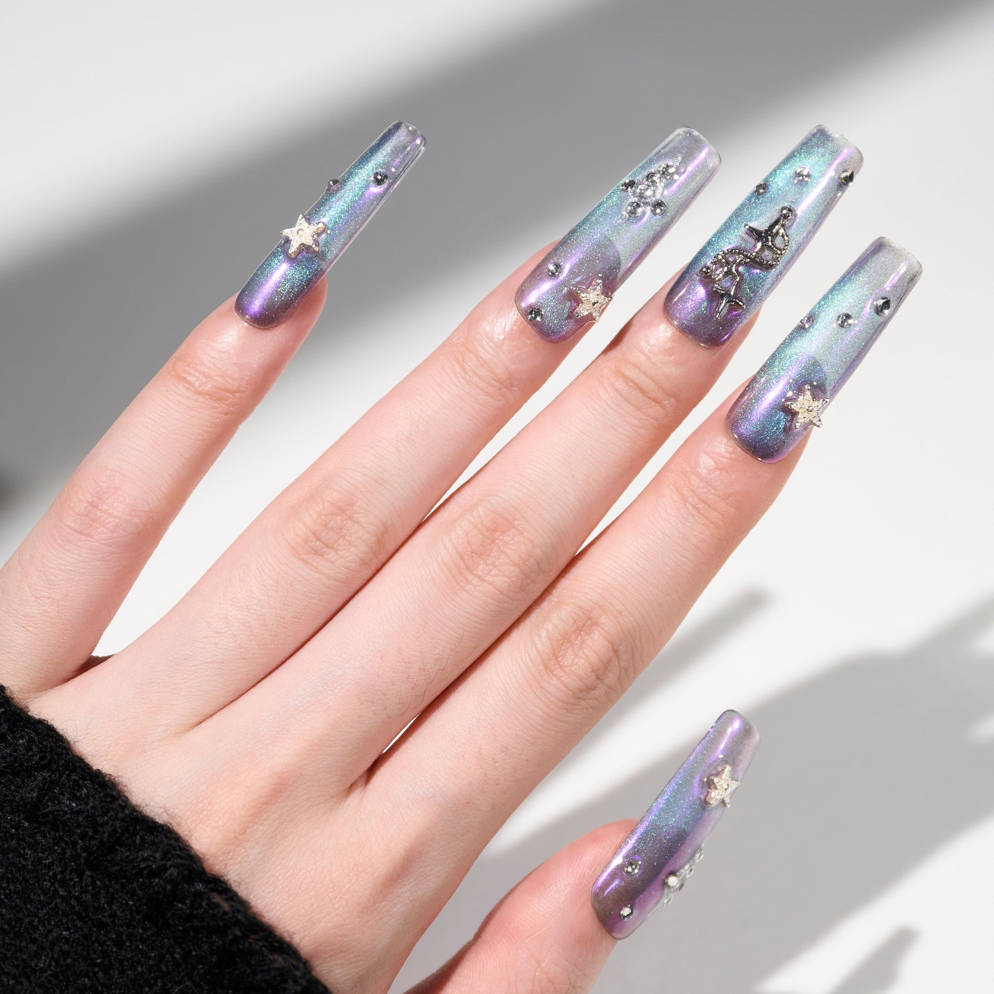 Hand with long iridescent blue-purple press-on nails decorated with rhinestones and star accents