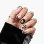 Hand with metallic silver and black press-on nails featuring star and cross designs, adorned with silver rings.