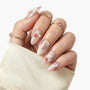 Hand with long almond-shaped press-on nails featuring pink shimmer, 3D floral designs, and tiny strawberry accents.
