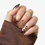 Hand with brown and gold almond-shaped press-on nails, featuring rhinestones and leaf designs