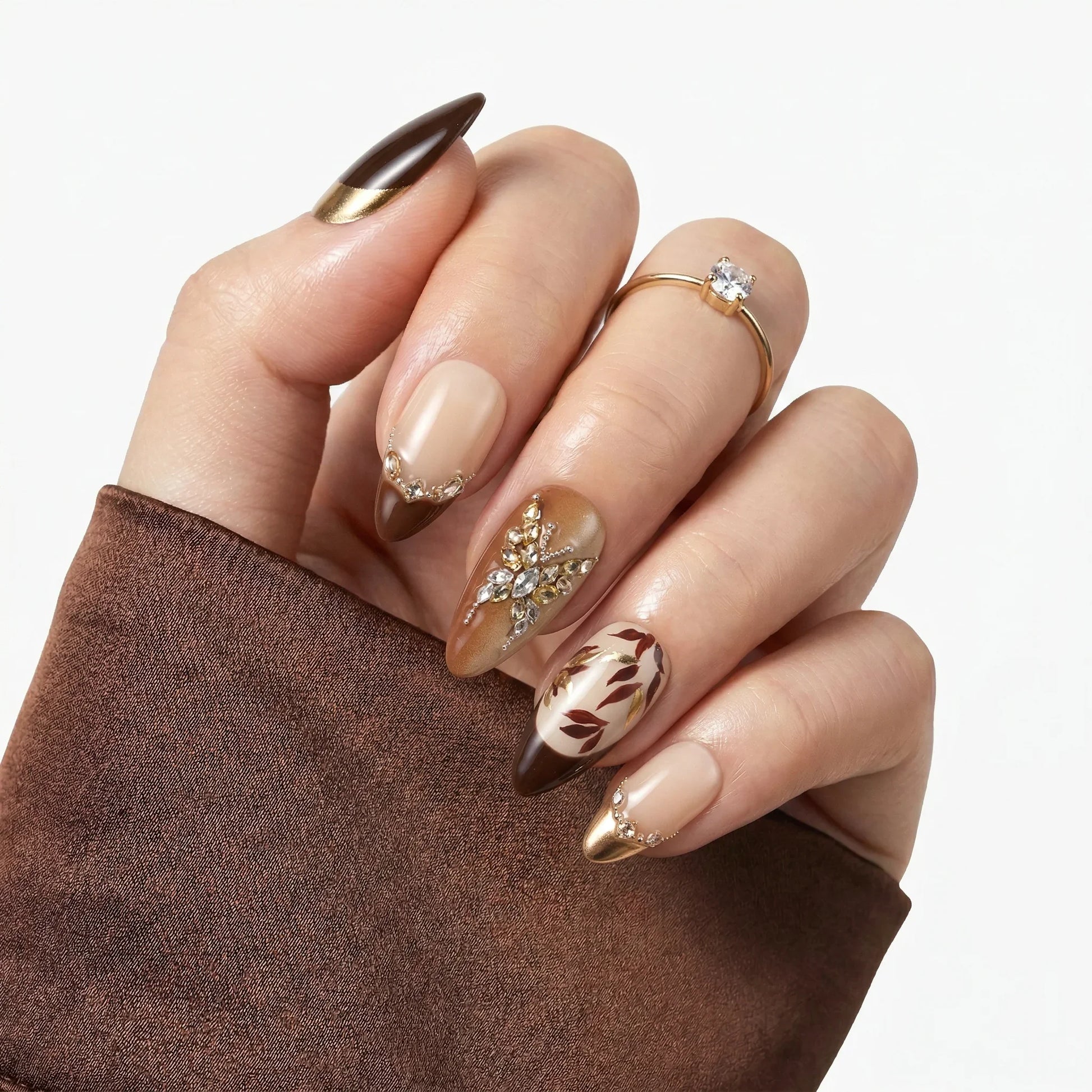 Hand with brown and gold almond-shaped press-on nails, featuring rhinestones and leaf designs
