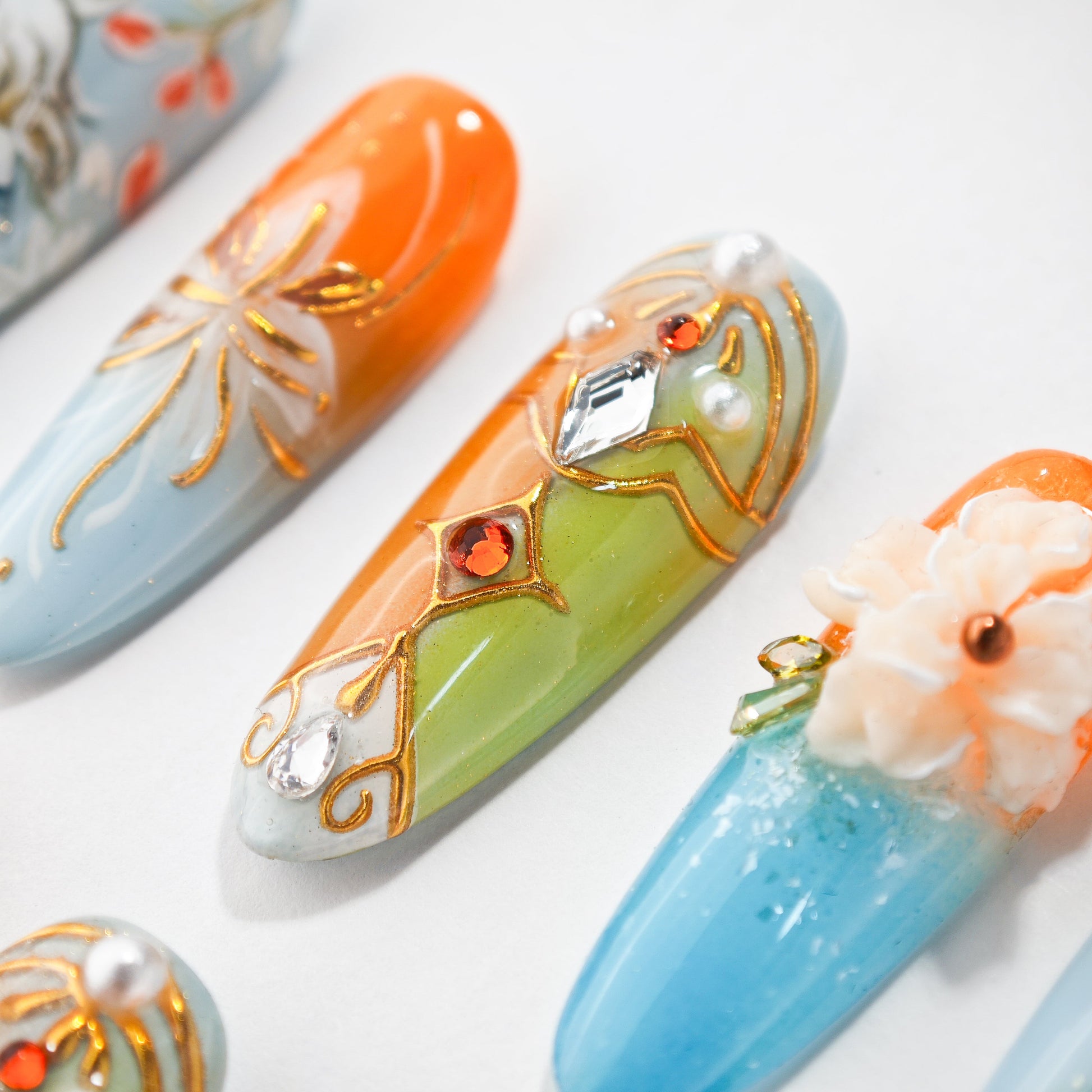 Colorful artificial nails with orange, green, and blue hues decorated with gold lines, gems, pearls, and 3D flowers