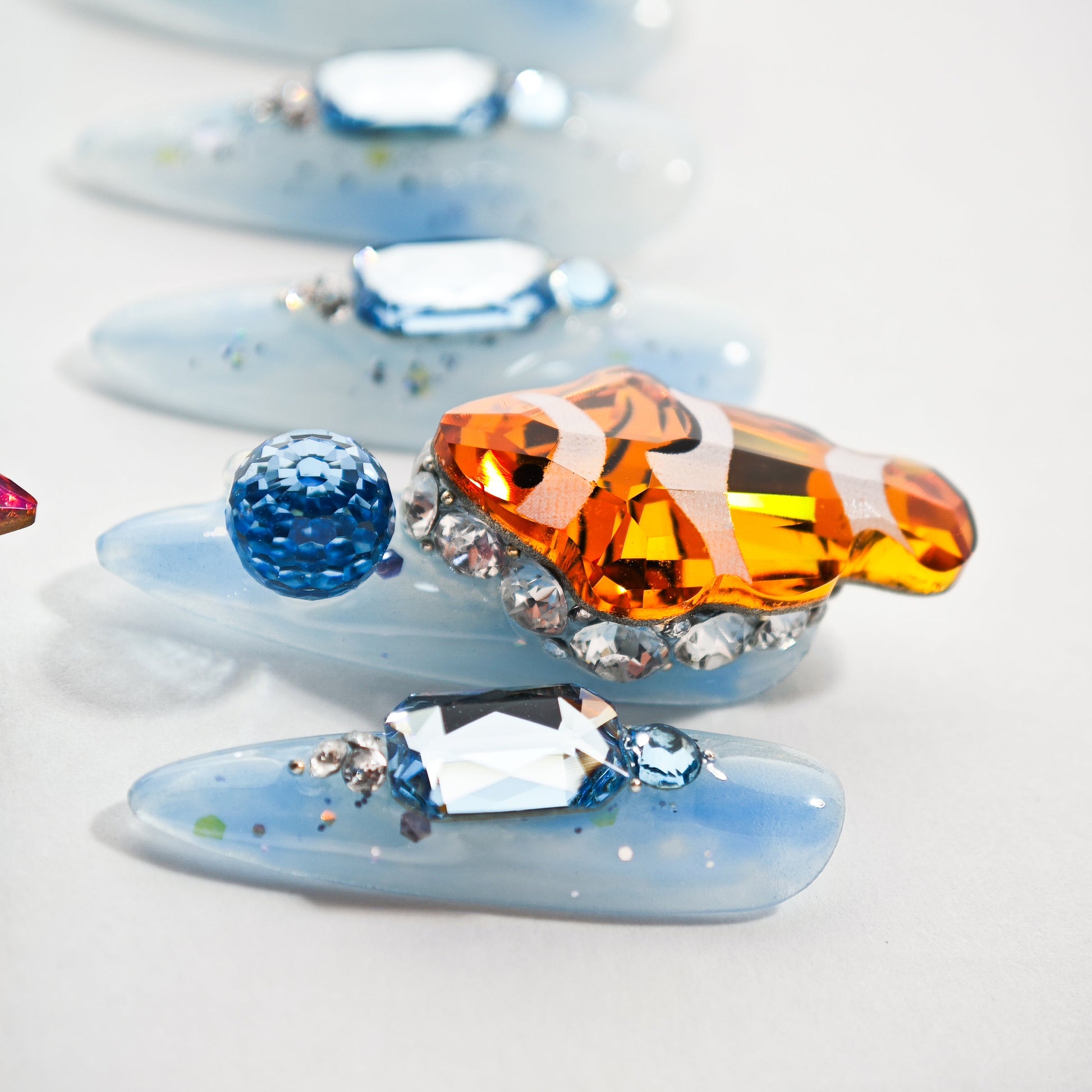 Blue acrylic nails with rhinestones and an orange crystal clownfish decoration on white background