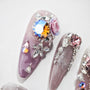 Handmade press-on nails with purple base, rhinestones, floral 3D design, and crystal gems