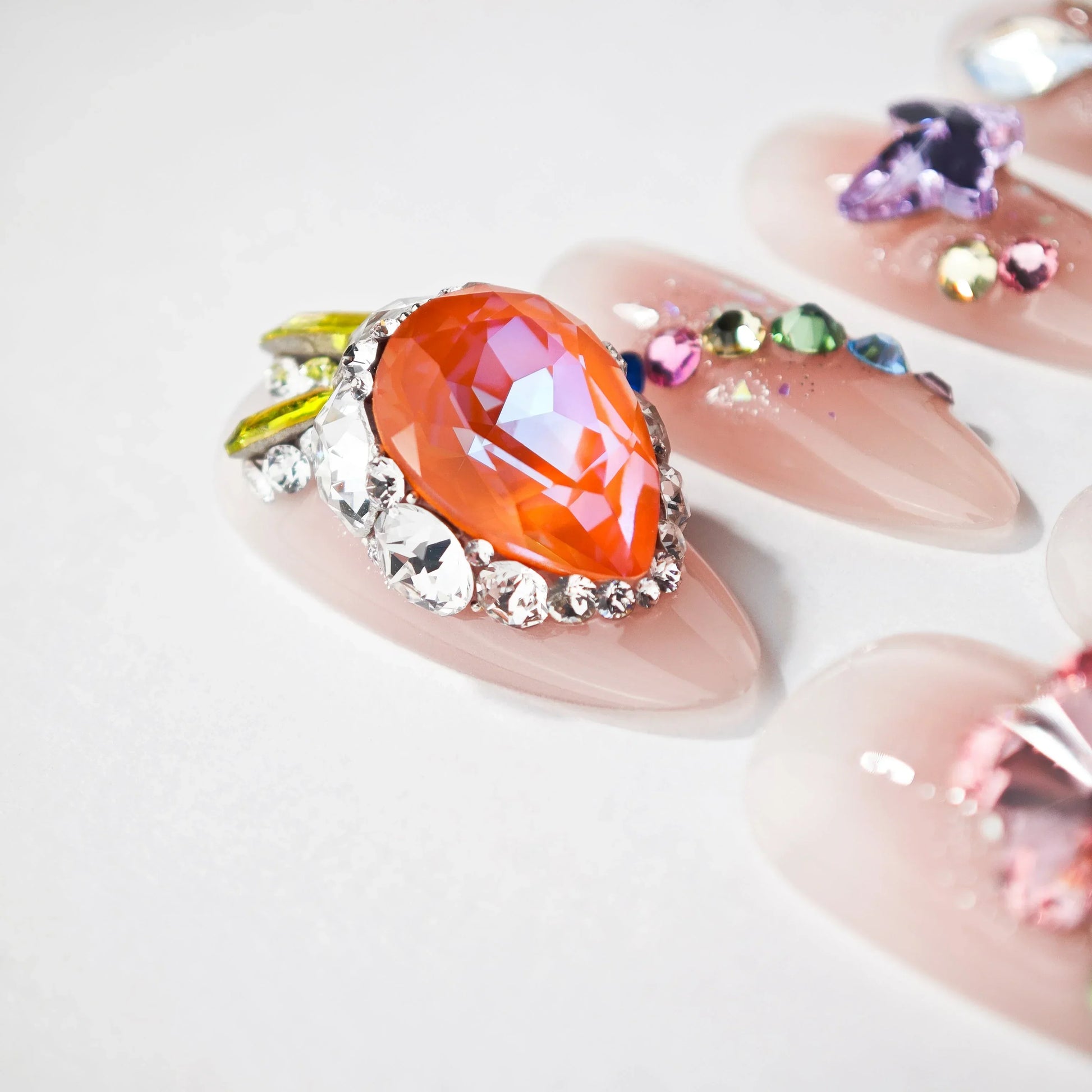 Nude pink artificial nails with large orange teardrop gem and multicolored rhinestone accents
